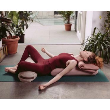 Kobieta w różowym stroju do jogi leżąca na macie w pozycji relaksacyjnej, wsparta na dwóch wałkach do jogi w odcieniach różu i  białym plażowym, z głową opartą na jednym z nich i zamkniętymi oczami. w tle rośliny i jasne wnętrze.